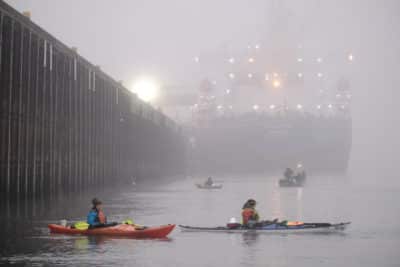 Activists in multicolored kayaks blockade a large barge--a pier is on the left side, and the sun is visible on the horizon through the gray mist.