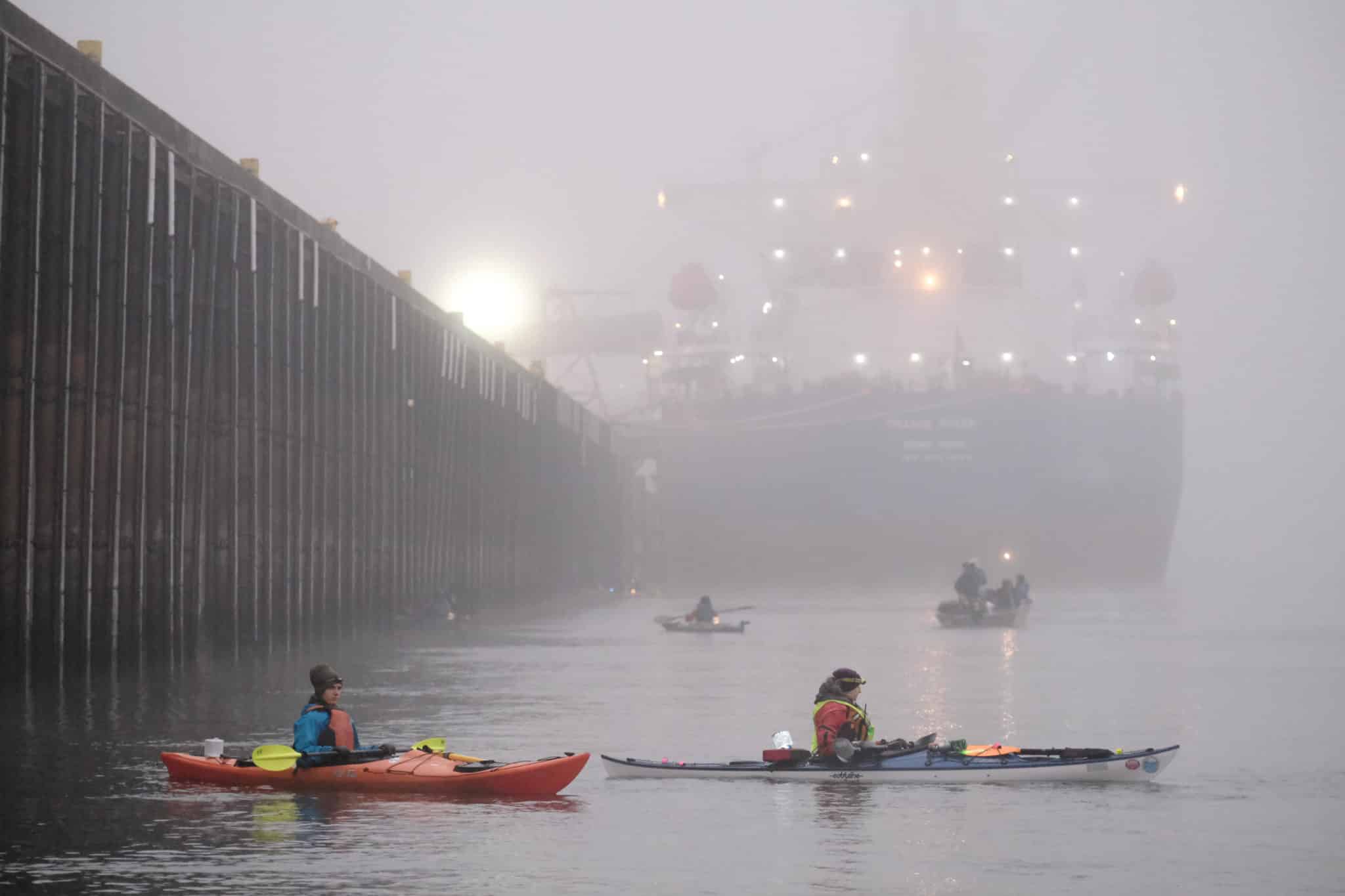 Activists in multicolored kayaks blockade a large barge--a pier is on the left side, and the sun is visible on the horizon through the gray mist.