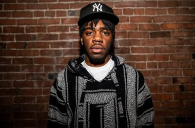 Portrait of Tyshawn Ford, wearing a black hat and hoodie and standing in front of a red brick wall.