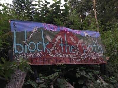 Hand painted banner hanging in forest reads: STOP THE MVP, BLOCK THE PATH, NO PIPELINE ON STOLEN LAND