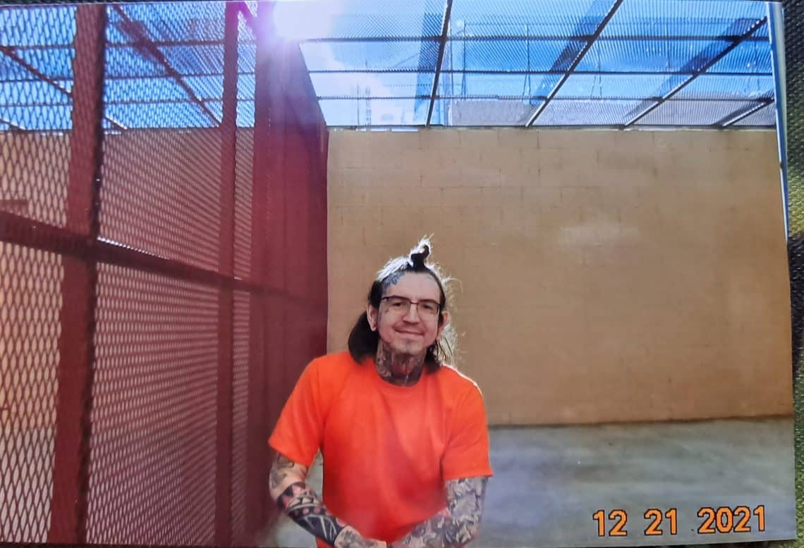 Eric King in an orange shirt, glasses, and smiling at the camera from within an outdoor prison cell.