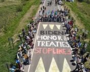 Water Protectors line a road blockade and the roadway has been painted with the words "BIDEN HONOR THE TREATIES: STOP LINE 3." The image is an aerial shot.