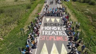 Water Protectors line a road blockade and the roadway has been painted with the words "BIDEN HONOR THE TREATIES: STOP LINE 3." The image is an aerial shot.