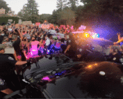 A group of protesters stand together with their hands up, masks on, bathed in the red and blue light of police vehicles.