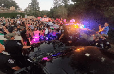 A group of protesters stand together with their hands up, masks on, bathed in the red and blue light of police vehicles.