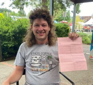 A man with curly brown hair and white complexion smiles while holding a pink parks citation. Text reads: City of Eugene Drops Charges Against Houseless Advocate