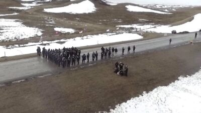 "Aerial view of a snowy landscape where a group of law enforcement officers stand in formation on a road, with some officers detaining individuals on the ground in the foreground. The area appears rural, with patches of snow covering the fields, and flags are seen lining the road in the background. The scene suggests a protest or confrontation in a remote setting."