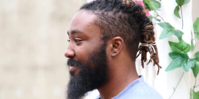 A side profile of a person with a beard and dreadlocks tied back, wearing a light-colored shirt. They are standing in front of a light, neutral background with some green foliage visible to the side, creating a natural and serene setting.