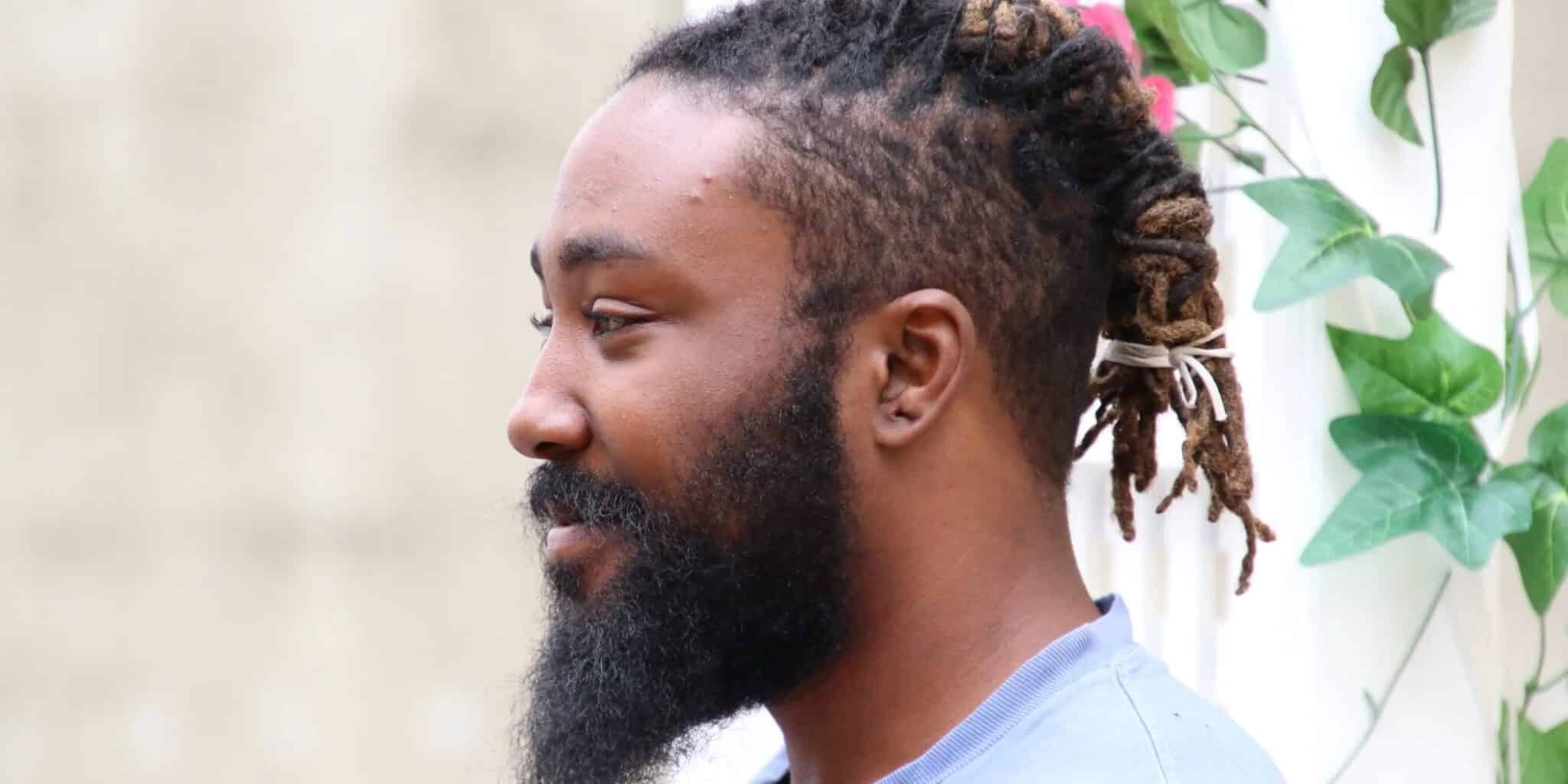 A side profile of a person with a beard and dreadlocks tied back, wearing a light-colored shirt. They are standing in front of a light, neutral background with some green foliage visible to the side, creating a natural and serene setting.