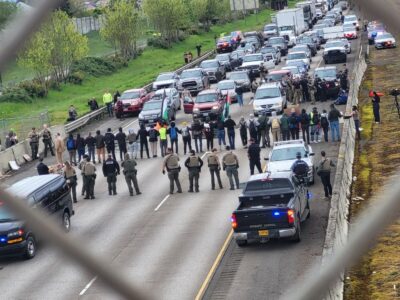 A group of protesters forms a human chain across a highway, blocking traffic in both directions. They are standing in a line, holding hands, and wearing backpacks and casual clothing. Several law enforcement officers, some in tactical gear, stand facing them. Police vehicles with flashing lights are positioned on the highway, and traffic is backed up behind the protest. Some officers appear to be making arrests or detaining individuals near the front of the blockade. A person is holding a Palestinian flag. The scene is viewed through a chain-link fence, adding a framing effect to the image.