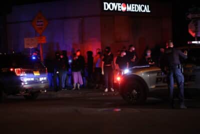 A nighttime protest outside Dove Medical, a facility with a brightly lit sign. A group of masked protesters dressed in dark clothing stands on the sidewalk, some holding signs. One sign reads 'F*** Your Violent Laws.' The scene is illuminated by the flashing red and blue lights of Eugene Police vehicles, which are blocking the street. A police officer stands near the vehicles, facing the protesters with one hand raised. The atmosphere appears tense, with the police presence and the demonstrators facing off in the dimly lit environment.