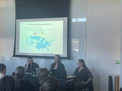 This image shows a panel discussion or presentation about "Critical Energy Infrastructure Laws" in what appears to be a conference room or lecture hall. There's a projection screen displaying a "US Protest Law Tracker" with a color-coded map of the United States showing different states in various shades of blue. The tracker appears to monitor legislation related to protest rights and energy infrastructure. In the foreground, three people are seated at a table facing an audience. From left to right: a person with glasses wearing a dark jacket with what appears to be a "RESIST" sticker on their laptop, a woman in dark clothing speaking (presumably presenting), and another person in a patterned dark top. There are audience members visible from behind in the foreground, and a trash can can be seen on the right side of the image. The room has white walls and the presentation appears to be discussing legal tracking of protest-related legislation across different states.
