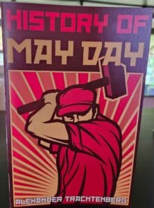 poster titled 'HISTORY OF MAY DAY' by Alexander Trachtenberg. The cover features Soviet-inspired graphic design with a purple background and red sunburst pattern. In the foreground is a stylized silhouette of a worker in a cap holding a hammer or tool over their shoulder. The title appears in bold letters with 'HISTORY OF' in red at the top and 'MAY DAY' in gold beneath it. The author's name is displayed at the bottom in white text.