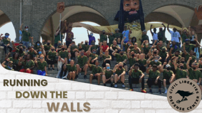 A large crowd of people, many wearing green shirts, sits in stadium-style seating with their fists raised in celebration. The setting appears to be under stone arches with brick architecture. A large inflatable figure with long dark hair looms above the crowd. In the foreground, there's text reading 'RUNNING DOWN THE WALLS' and a circular logo for 'Civil Liberties Defense Center' featuring a raven and keys.
