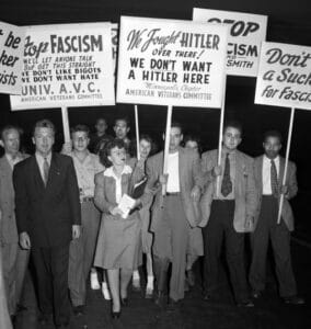 Here's alt text for this historical photograph: Alt text: A black and white photograph showing a group of protesters in 1940s attire holding signs with anti-fascist messages. The signs include text such as "Don't be a sucker for FASCISM," "We fought HITLER over there! WE DON'T WANT A HITLER HERE," and references to the American Veterans Committee. The protesters, dressed in suits and formal wear typical of the 1940s era, appear to be participating in a demonstration against fascism and hate groups in America. This appears to be documentation of post-World War II anti-fascist activism in the United States, when veterans and citizens organized to oppose the spread of fascist ideologies domestically after fighting against them abroad.