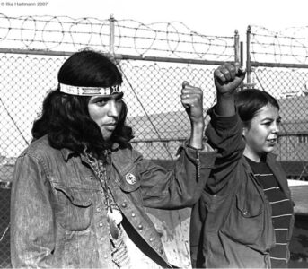 Two young Indigenous activists raise their fists in solidarity at a protest. They stand in front of chain-link fencing topped with barbed wire. The person on the left wears a headband and denim jacket with pins, while the person on the right wears a striped long-sleeve shirt. The black and white photograph captures a moment of youth activism and resistance. Photo credit: Ilka Hartmann 2007