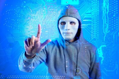 A figure wearing a white featureless mask and gray hooded sweatshirt reaches toward the viewer with their right hand against a bright blue digital background filled with circuit board patterns, binary code, and glowing technological elements. The person's hand appears to be interacting with a translucent holographic interface, creating a cyberpunk aesthetic that suggests themes of digital anonymity, hacking, or technological interaction.