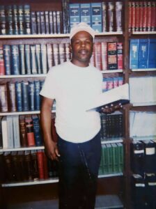 Calvin Duncan stands in front of floor-to-ceiling bookshelves filled with legal volumes at the Angola prison law library. Wearing a white T-shirt, white cap, and dark pants, he holds an open book in one hand while looking directly at the camera.