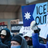 Bundled-up protesters at an anti-ICE demonstration in downtown Minneapolis hold signs demanding an end to immigration enforcement. In the foreground, a protester wearing a blue winter coat, knit hat and face covering raises a large blue “ICE OUT” sign featuring a white star. Other demonstrators carry signs reading “ABOLISH ICE” and “ICE OUT.”