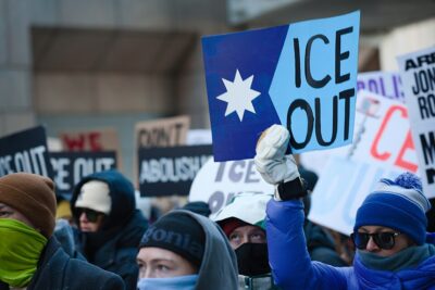 Bundled-up protesters at an anti-ICE demonstration in downtown Minneapolis hold signs demanding an end to immigration enforcement. In the foreground, a protester wearing a blue winter coat, knit hat and face covering raises a large blue “ICE OUT” sign featuring a white star. Other demonstrators carry signs reading “ABOLISH ICE” and “ICE OUT.”